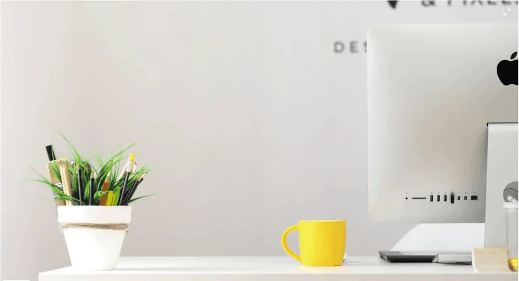 A desk with a computer, plant and yellow coffee mug on it.