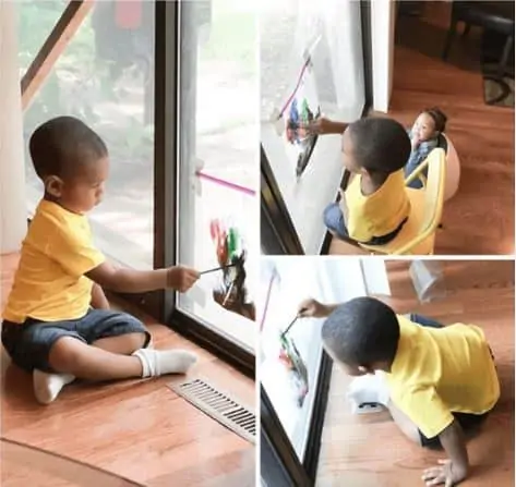 A young boy painting on the window with window paints.