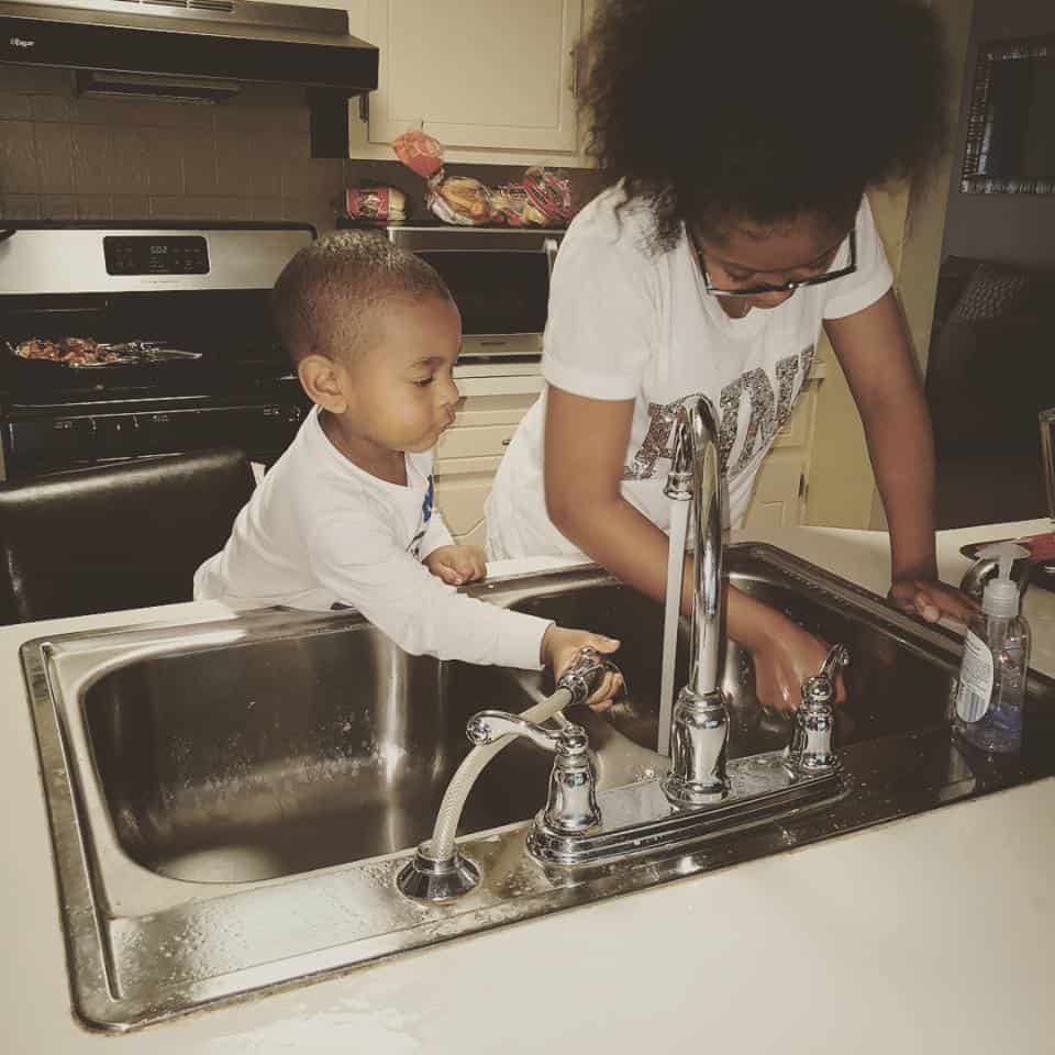 Teen working as a babysitter and washing dishes with younger child.