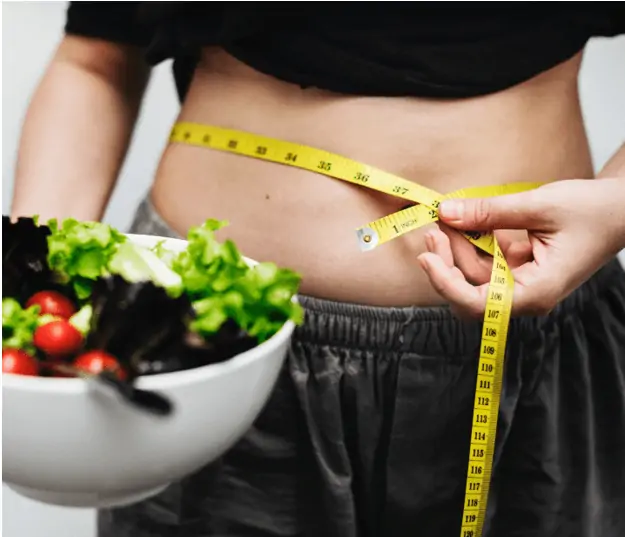 A woman that is participating in a weight lose challenge is measuring her waist and holding a salad