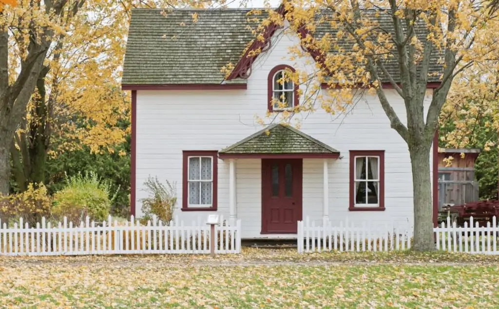 White home with red trim and white fence in the woods.