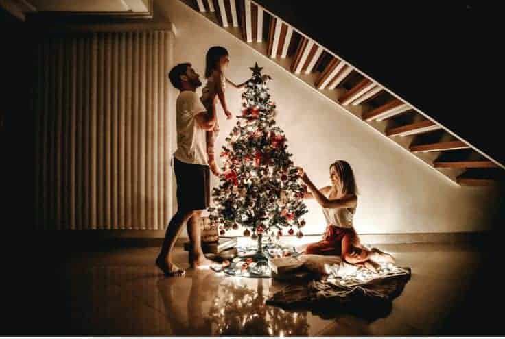A man and woman decorating a christmas tree with their little girl.  The dad is holding the little girl up to put the star on top